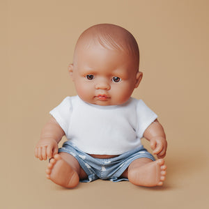 Miniland Baby Puppe mit brauner Haut, weißem T-Shirt und blauen Shorts sitzend