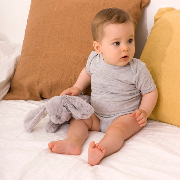 Ein Baby sitzt auf einem Bett mit einem grau Plüsch-Baby-Projektor und schaut neugierig zur Seite