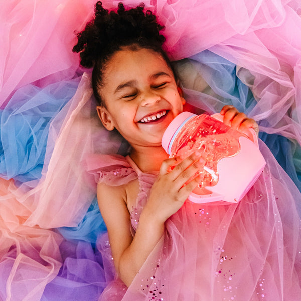 Kind hält lachend Sensorikglas zum Befüllen mit Wasser – rosa von Glo Pals in buntem Tüllkleid