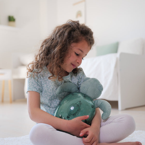 Mädchen mit Tranquil Turtle Spieluhr Projektor in der Hand, sitzt auf dem Boden in einem hellen Raum.